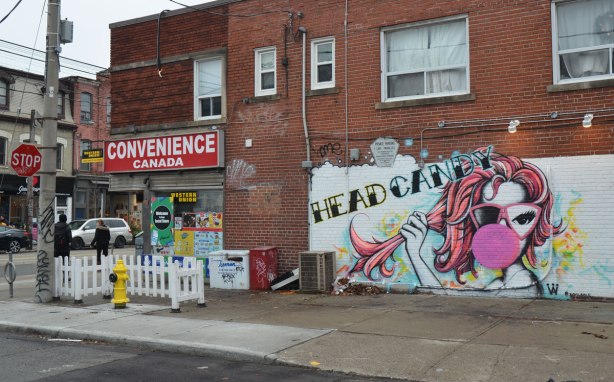 on the corner of Queen West and a smaller side street, the Convenience Canada store with a small white picket fence outside of it. On the wall is a mural of a woman blowing a big pink bubble with gum and the words head candy written above her head.