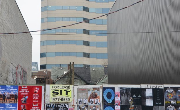 three layers, in the background a high rise building, in the middle ground, the tops of three single family homes on Richmond Street, in the foreground, poster covered hoardings for construction on Queen Stree West.