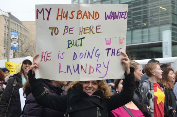a woman holds a sign up above her head. It says My husband wanted to be here but he is doing the laundry 