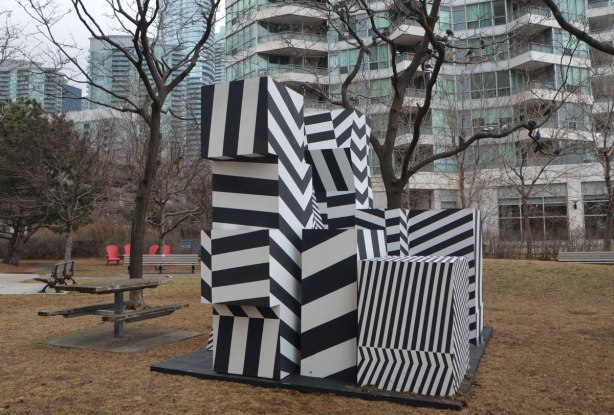 a public art installation in a park, winter time but no snow, no leaves on the trees, muddy brown grass, condo in the background. Artwork is a series of shapes stacked in a pile, all shapes are painted in black and white wide stripes. The stripes are horizontal, vertical and diagonal in the resulting structure, called incognito, by Jaspal Riyait and Curio