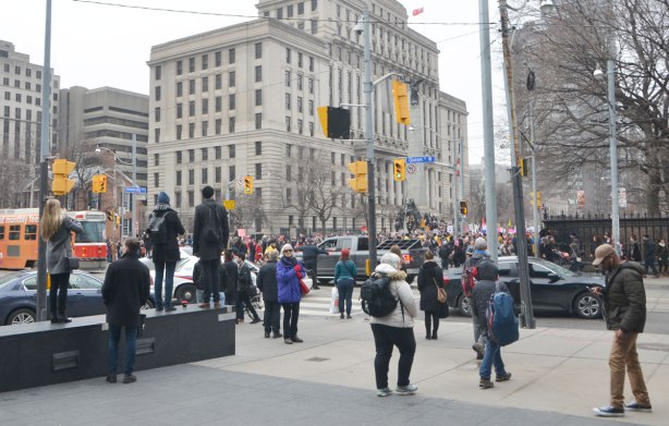 intersection of Queen and University, people standing on the sidewalk looking at the Womens March as it stops on University. Policemen trying to direct traffic on Queen as they prepare to close Queen Street for the march 