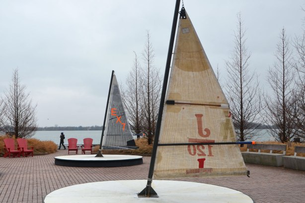 with the lake in the background, art installation beside Lake Ontario on Toronto's waterfront, called Leeward Fleet, by RAW consulting, two (of three) round platforms with a small sail in the middle. The boom of the sail is the handle for turning the platforms round and round like a merry go round. 