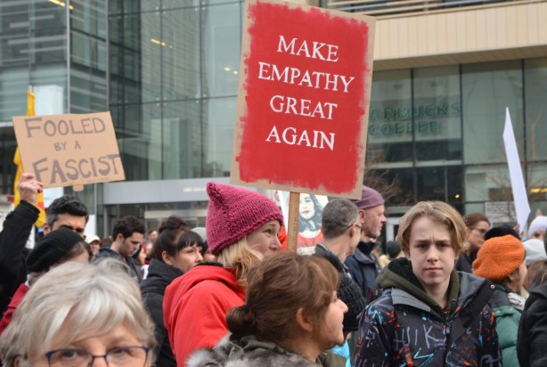 a woman walking in the Womens March, toronto holds up a read sign that says Make Empathy great again. Lots of other men and women walking in the same picture. 