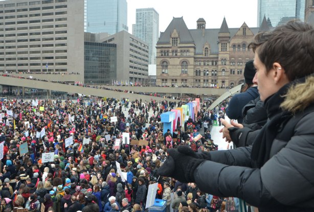 Taken from the upper level at Nathan Phillips Square, overlooking the square which is full of people attending Womens March, toronto . In the foreground are a couple of people who are also on the upper level. 
