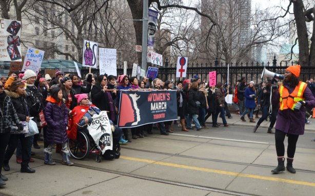 Womens March, toronto - the head of the march goes past Osgoode Hall on QUeen Street. A woman with a megaphone is leading the chants and singing. A large group with a banner that says Womens March is the first group in the walk 