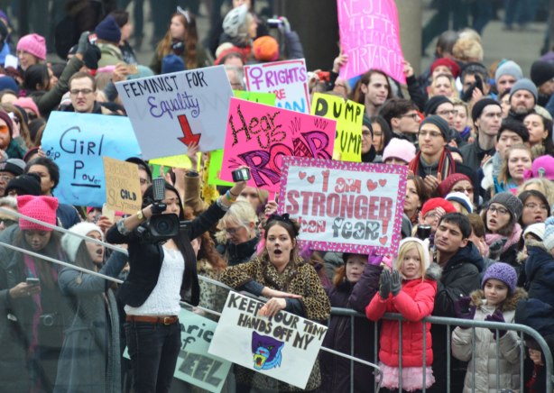 standing behind the barricades by the stage at Nathan Phillips Square, a large group of people at the Womens March, toronto . Many signs and many people. and a reporter with a camera.