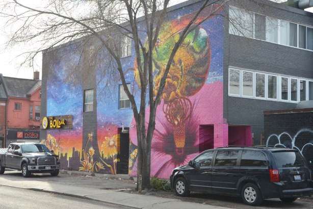 large mural of a honey bee on a big pink flower hibiscus, with other yellow flowers growing, also a black skyline of Toronto, a large tee grows in front of the building and a van and a truck are parked close by. Photo taken from across the street. 