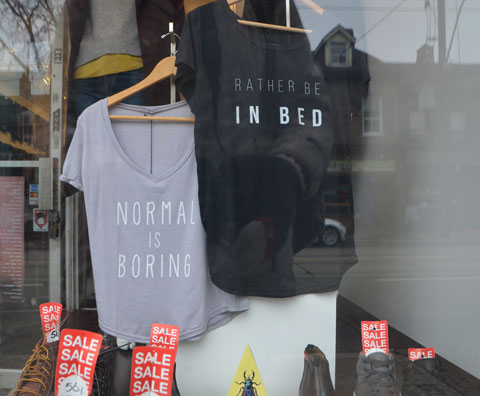 Two t-shhirts in a store window, a grey woman's t-shirt with the words Normal is Boring on the front and a black t-shirt with the words rather be in bed.
