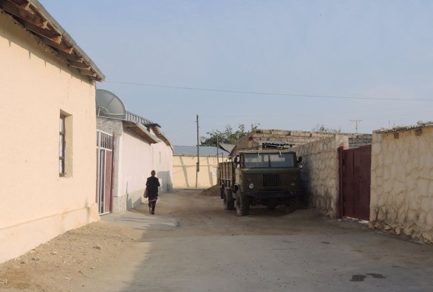 street in Naruta Uzbekistan with yellowish coloured mud walls lining the street - a woman walks up one side of the street, past a green truck parked on the other side of the street