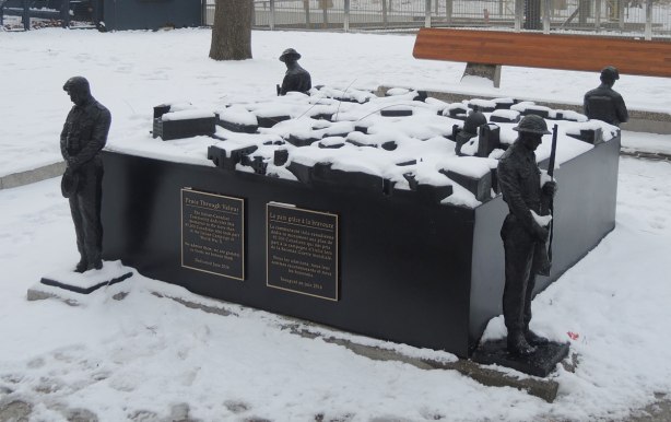 a sculpture called Peace Through valour by Ken Lum, outside on a snowy day. A square piece with a soldier standing guard at each corner. On top of the flat squsre is a model of a town in square blocks (no details on the buildings). 