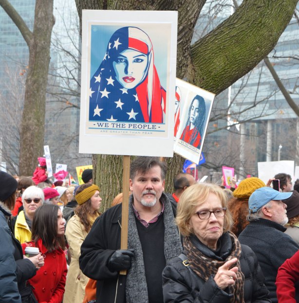 a man holds a sign up in the air, above the heads of out protesters at the Womens March, a Shepard Fairey design of a woman in stars and stripes American flag head scarf withthe words "We the People"