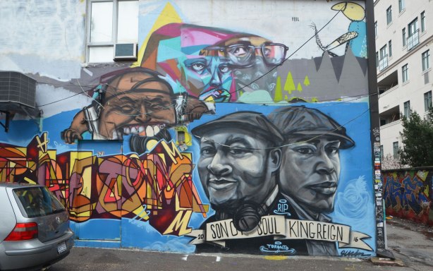a wall in Graffiti Alley, layers of different artist's work, some uber 5000, some elicser, plus a grey ton picture of two men wearing caps with the words son of soul king reign written underneath them.
