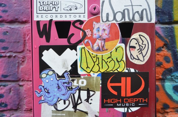 stickers on a small metal box on a brightly coloured wall. almost the whole box is covered, a lovebot heart, a pink cat, a blue octopus, a shark, plus some stickers that are ads for music or records.