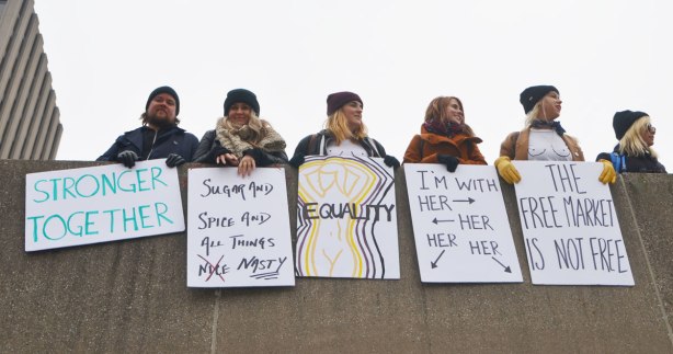 Womens March, toronto - a group of people leaning over the edge of the upper level at Nathan Phillips, holding their placards and signs over the concrete. Two of the women are wearing white T-shirts on which they've drawn black outlines of breasts. 