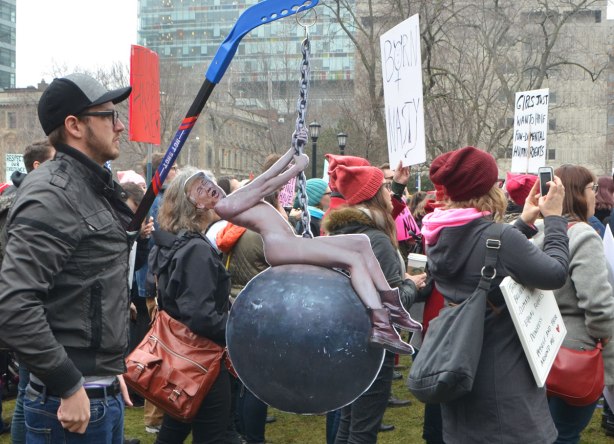 protest march and rally at Queens Park, large crowd of people with signs and pink hats. A man has a cutout of a naked Donald Trump swinging on a large wrecking ball. 