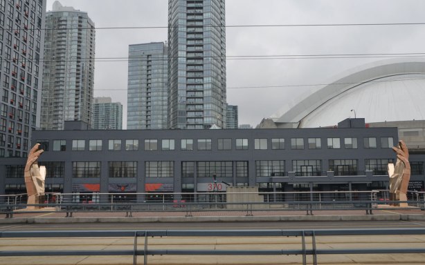 looking across Queens Quay west to the buildings and condos in downtown, the edge of the Skydome (Rogers Center) is just visible, two large hands rise upwards as part of an art installation. 