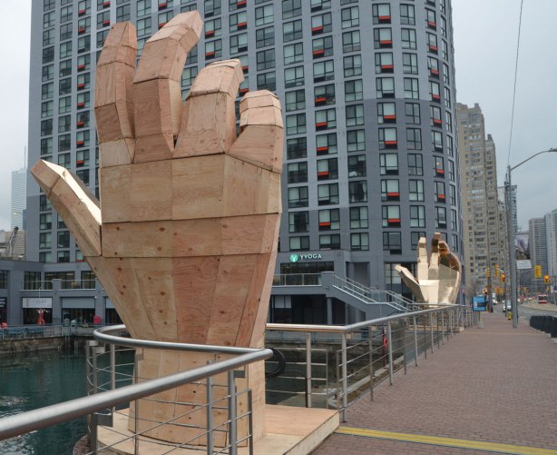 two large wooden hands face each other, beside the sidewalk, condos behind, the plams are covered with shiny gold, 