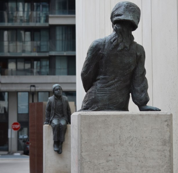 two children of toronto, a sculpture by Ken Lum, two children seated on pedestals, about 25 feet apart, along the side of a walkay, with a concrete building beside them. The children are looking towards each other 