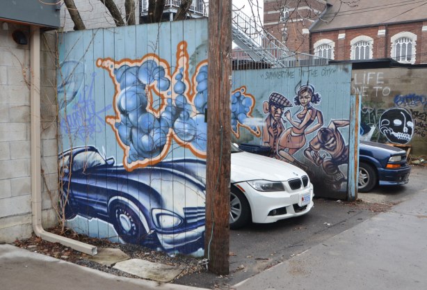 in an alley, a white car is parked between a blue car and a mural painting of a blue car. There is also a mural of three people with the words summertime in the city.