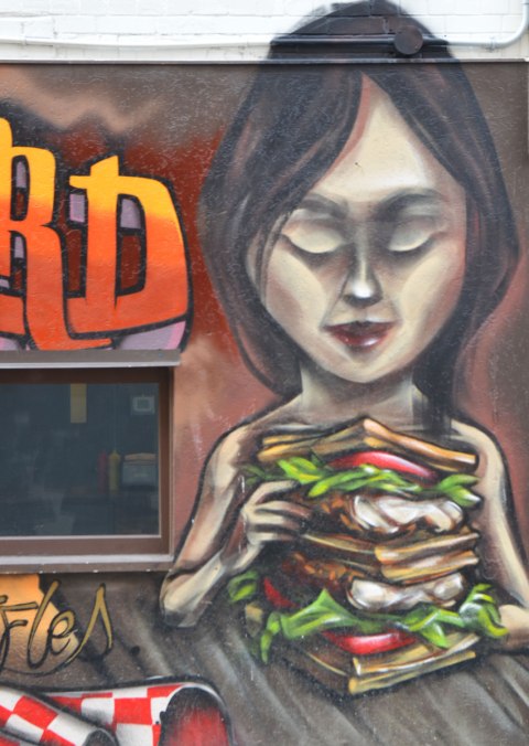 from a mural by elicser along the exterior wall of the dirty bird chicken and waffle restaurant - a woman is sitting at a table in the restaurant with a very tall chicken and waffle sandwich in her hands