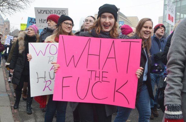 a young woman amidst a crowd of others walking in the Womens March in Toronto, holding a large pink sign that syas what the fuck 
