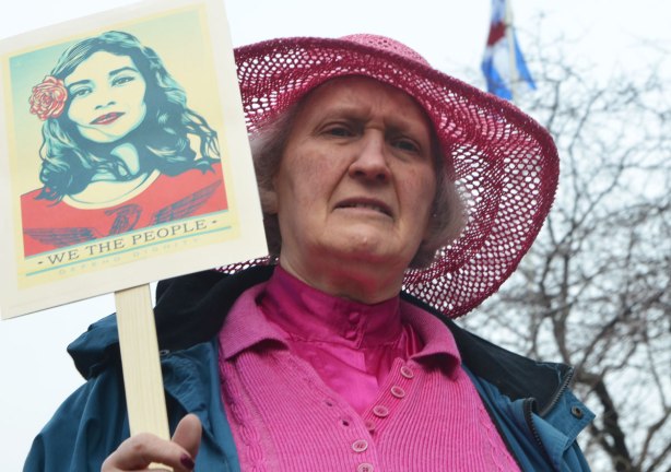 an older womaloosely woven pink hat with wide brim, and a pink top, holds a sign at a march 