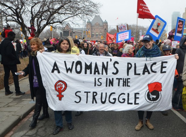 a group holds a white banner as they walk in the Womens March, the banner says A woman's place is in the struggle. 