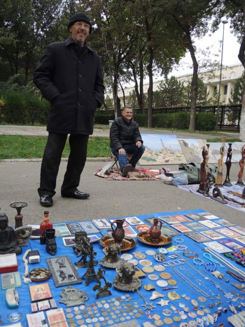 two men are selling items on the sidewalk. They have the things laid out on a blue piece of fabric