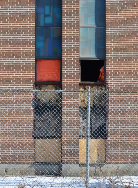 vertical windows on a birck church, behind a chain link fence. Two of the windows have panes missing and are covered with orange cloth 