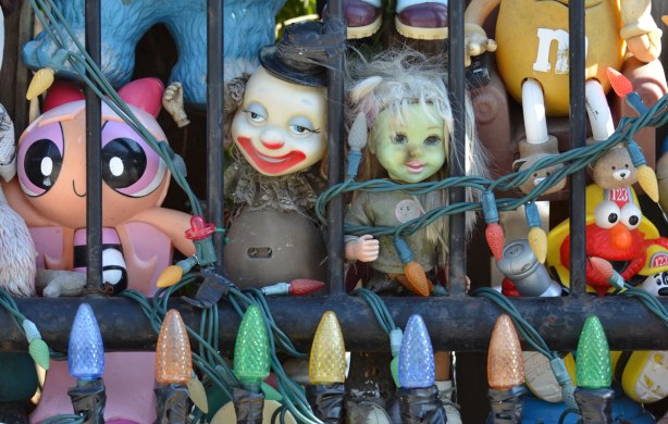 peering between the metal bars of a fence is a creepy clown doll and a green faced zombie doll, a string of Christmas lights is across the bottom of the fence 