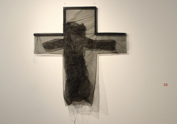 black netting in the shape of a cross on a wall. within the cross is more black light weight fabric in the shape of a person whose arms in the horizontal parts of the cross. 