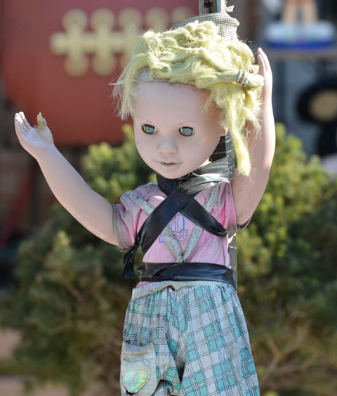 a faded blond doll with blank eyes looks down, she is attached to a wooden stake with black electrical tape 