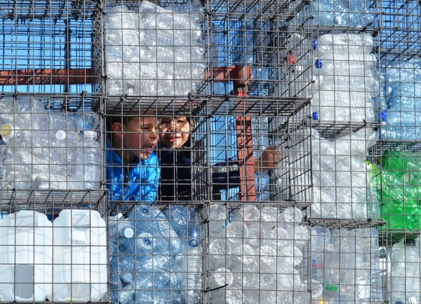two boys peer out from behind a wall of wire cages filled with empty plastic bottles. One of the cages is empty as looks like a window 