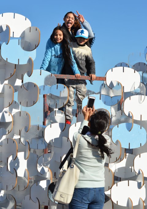 three kids stand on top of a lifeguard station that is enclosed by an art installation that is construction of many oval shapes joined together. Some are white, some are clear and some are reflective. A mother is taking a picture of the kids. 