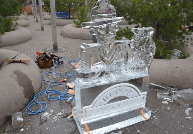 an ice sculpture of a mountie and a beaver has just been finished. The tools used by the artist are lying around on the ground below the sculpture. 