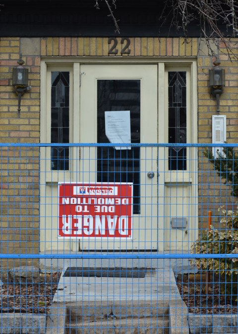 the front door of a small apartment complex that is about to be demolished. There is a blue metal fence in front of it with a danger due to demolition sign on it. The sign is upside down. 