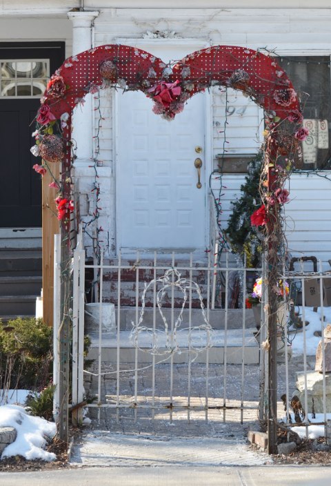 a small archway over a gate at the entrance to a front yard, the arch is the shape of a heart and it has been decorated with flowers. 