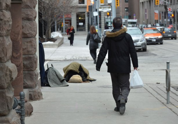 people walk by on the sidewalk as a homeless man sleeps under blankets on the corner. 