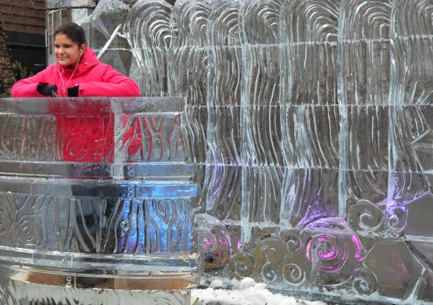a girl in a bright pink jacket stands behind a podium built of ice and in front of a wall made of ice. Both have curvy lines and swirls carved into them. 