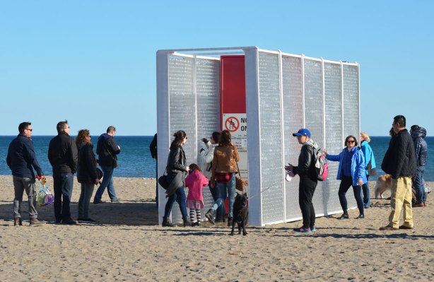 an art installation on the beach, people in winter clothes, two parallel walls about 10 feet high made of a layer of horizontally arranged empty plastic bottles with the opening facing in, people are writing on paper and then putting the messages in the bottles. 