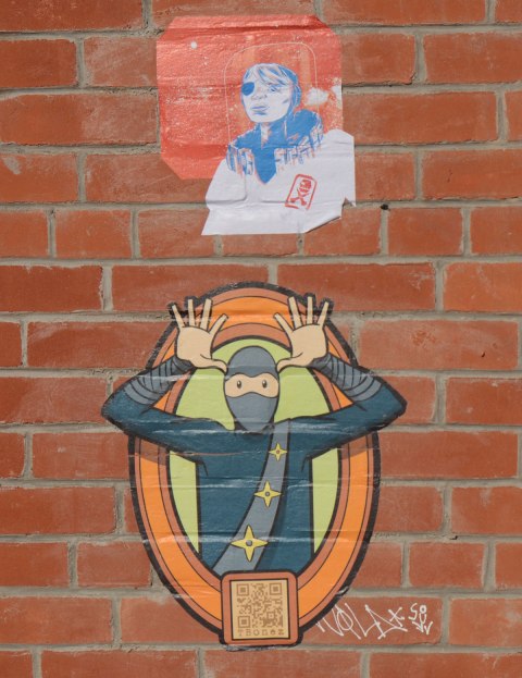 two paste ups on a red brick wall. the lover one is a tbonez character in his blue suit and grey mask, with hands beside face like going na na na na, can't catch me. or else hands look like moose antlers. upper paste up is a person's head. signature on upper one is like a skull over cross bones. 