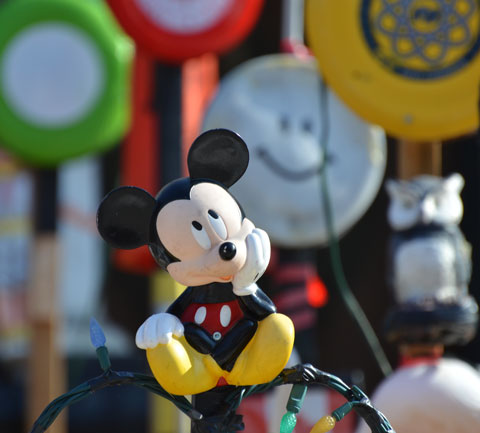 a mickey mouse plastic figure is sitting on a wire fence, his chin is his hand and he's looking upwards, other toys out of focus behind him - circles with happy faces and a couple of frisbees