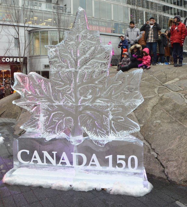 a group of people sit and stand on a large rock behind an ice sculpture of a maple leaf with the words Canada 150 under it, all carved in ice. 