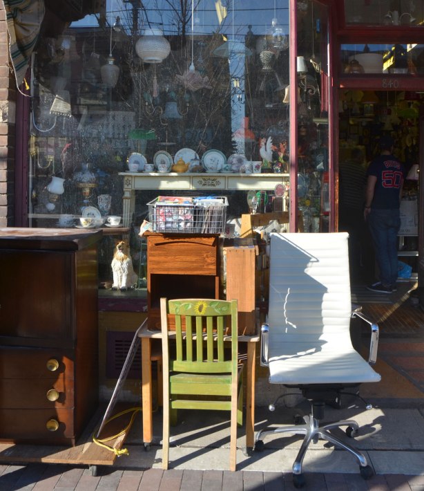 chairs and a set of drawers outside a store full of antiques and stuff, the door is open and you can see into the store where there are two men shopping. 