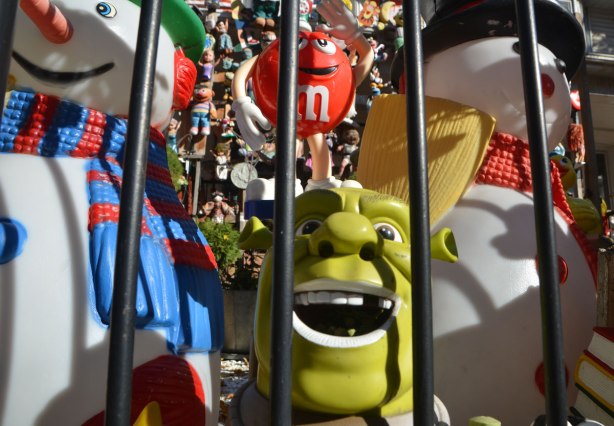 Shrek is between two snowmen, all plastic toys and decorations, behind the metal bars of a fence, a lot of toys and dolls and stuffed animals behind them. 