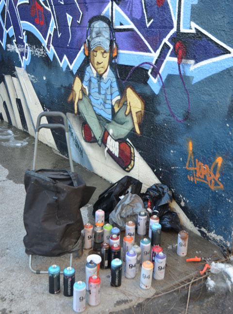 part of a mural of a boy with headphones on, seat crossed legged on the ground and pointing downwards. He seems to be pointing at a collection of spray paint cans sitting on the ground below the mural. 