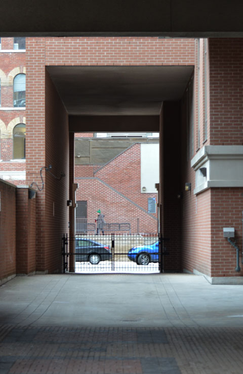 looking down a driveway that passes under a very high square arch to the street beyond. Cars are parked on the street and a pedestrian walks by 