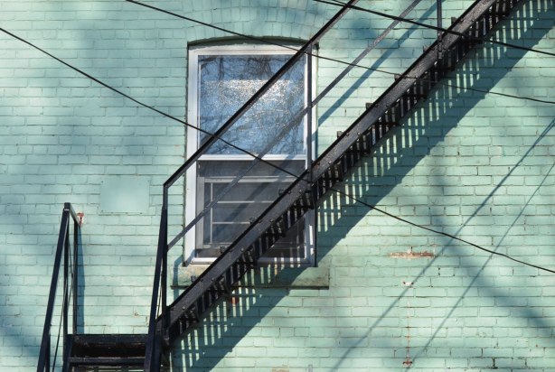 shadows on a sunny day, a metal fire escape is diagonal across the back of a light teal coloured house, it passes the bottom corner of a window 