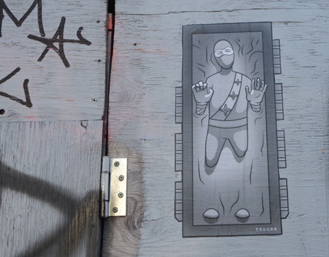 a monotone grey paste-up by tbonez on a grey garage door. Vertical, tbonez guy is lying on his back in wet concrete with hands and knees up or else he has run into a window. 