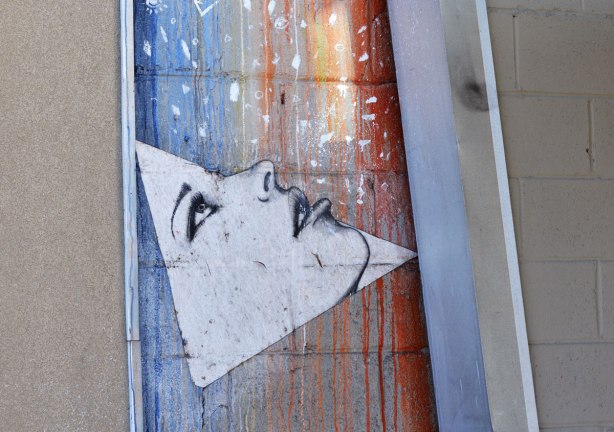 street art picture of a triangular shaped face looking upwards, open eyes and partially open mouth, behind the face is streaks of blue, yellow and red. 